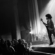 Black and white photo of a concert shot from the right side of the stage. The left half of the image is darkened with the audience on the bottom left. Light streams in from the top right down to the middle center, illuminating the stage. The sihouette of a guitar player can be seen at the front of the stage, approaching the crowd.