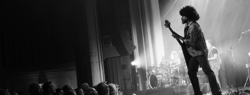 Black and white photo of a concert shot from the right side of the stage. The left half of the image is darkened with the audience on the bottom left. Light streams in from the top right down to the middle center, illuminating the stage. The sihouette of a guitar player can be seen at the front of the stage, approaching the crowd.