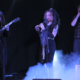 Photo of 3 muscicians performing against a black background with blue light on them. Smoke is coming in from the lower right to the middle of the image.