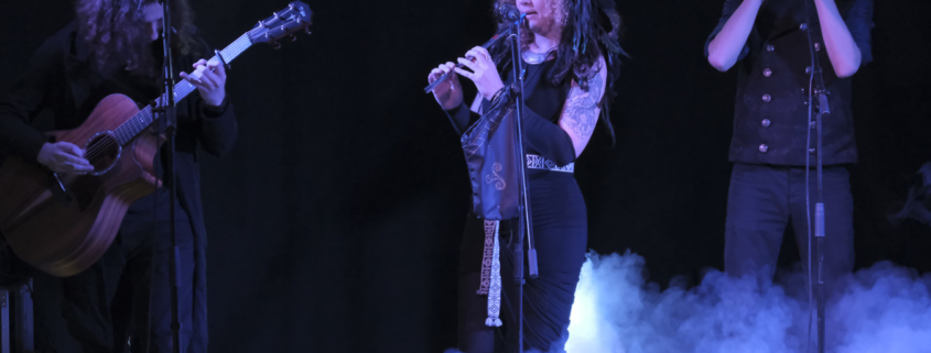 Photo of 3 muscicians performing against a black background with blue light on them. Smoke is coming in from the lower right to the middle of the image.