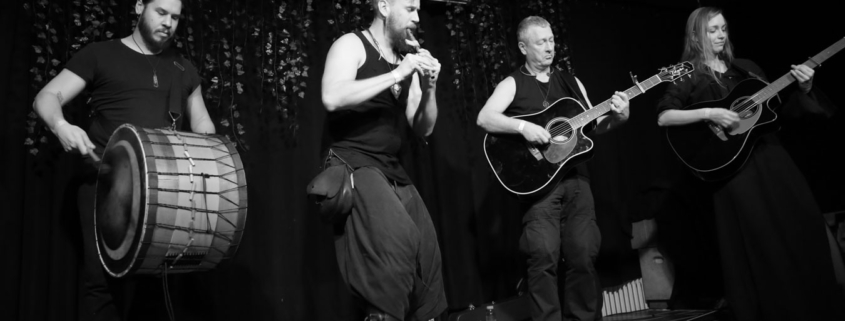 Black and white photo of the band Seed performing. From left to right: a man playing Davul drum, a man playing whistles, a man playing guitar and a woman playing bouzouki