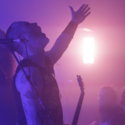 photo_2024-03-15_Skalmold Photo of 2 members of the band Skálmöld on the left and some adience members on the right bottom with a pinkish light in the background. One of the band members has their arm raised, fingers wide in the air while singing