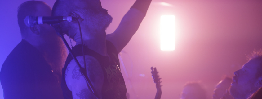 Photo of 2 members of the band Skálmöld on the left and some adience members on the right bottom with a pinkish light in the background. One of the band members has their arm raised, fingers wide in the air while singing